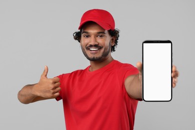 Happy courier holding smartphone with empty screen and showing thumbs up on light grey background