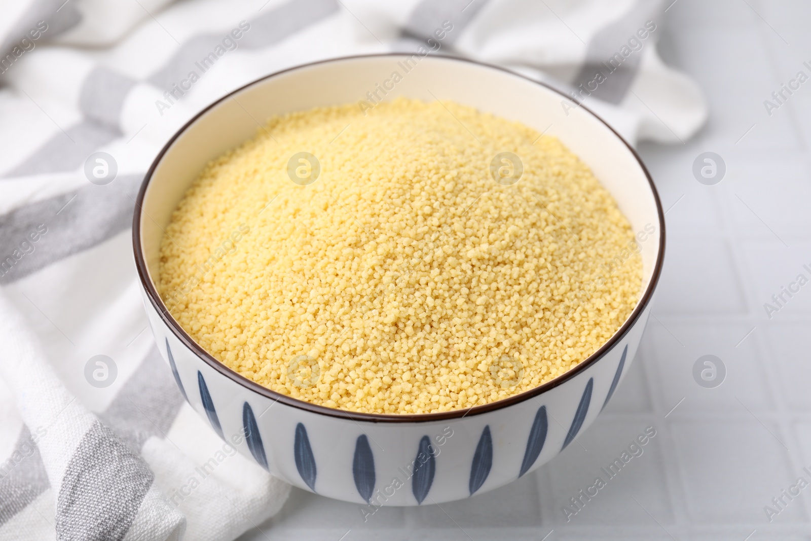 Photo of Raw couscous in bowl on white tiled table, closeup