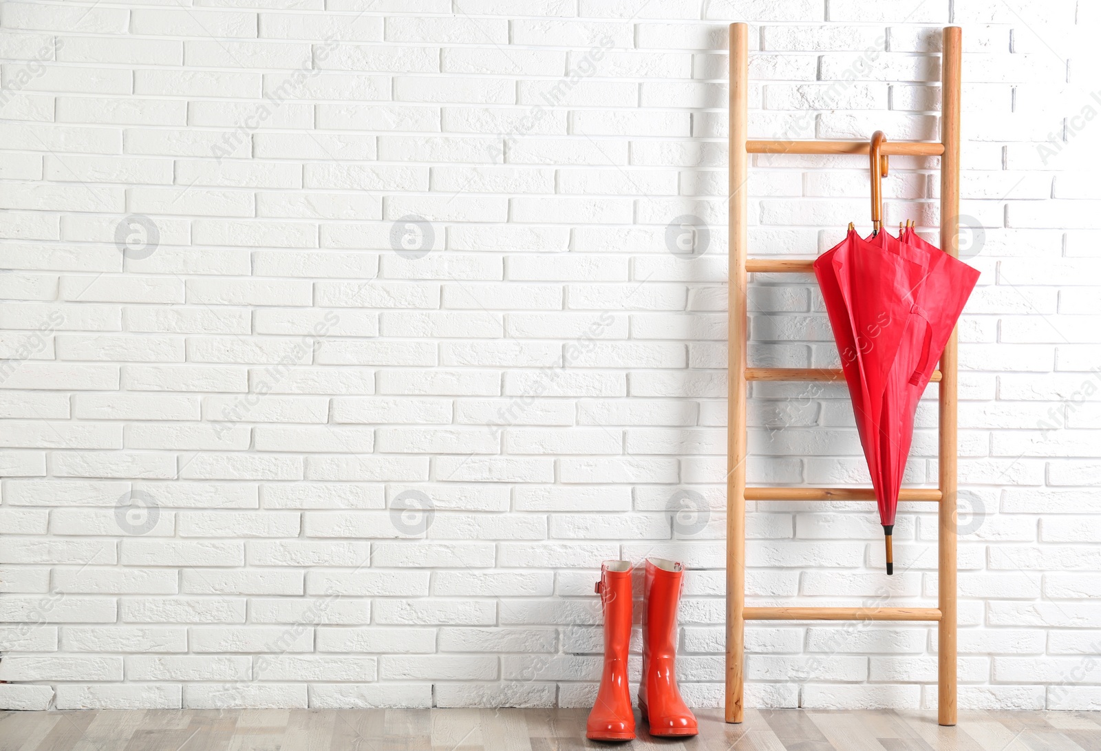 Photo of Beautiful red umbrella on ladder and rubber boots near white brick wall. Space for text