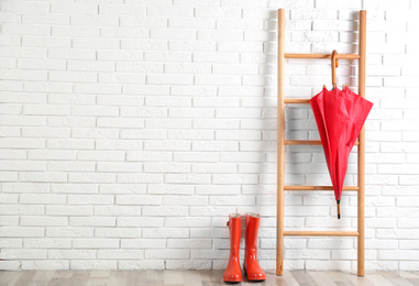 Beautiful red umbrella on ladder and rubber boots near white brick wall. Space for text