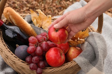 Woman taking apple from wicker picnic basket outdoors on autumn day, closeup