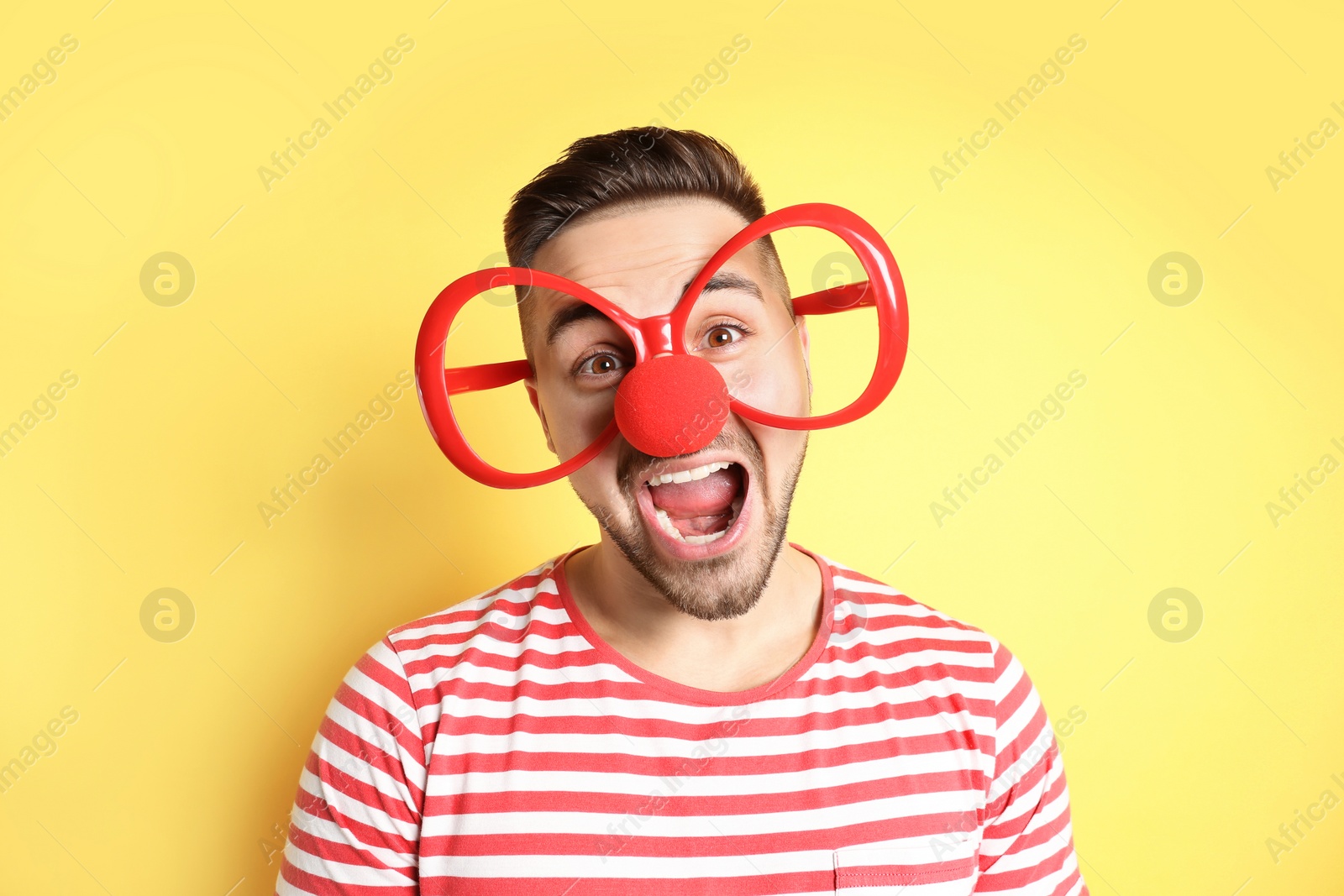 Photo of Emotional young man with party glasses and clown nose on yellow background. April fool's day