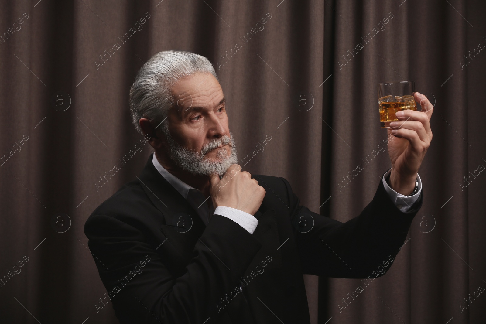 Photo of Senior man in formal suit holding glass of whiskey with ice cubes on brown background