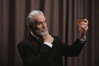 Photo of Senior man in formal suit holding glass of whiskey with ice cubes on brown background