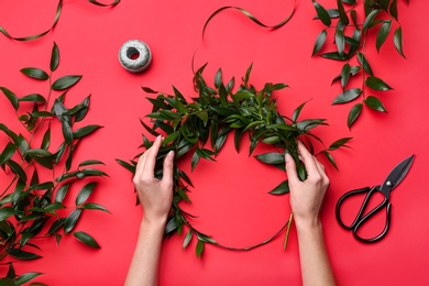 Photo of Florist making beautiful mistletoe wreath on red background, top view. Traditional Christmas decor