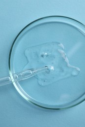 Photo of Petri dish with sample of cosmetic oil and pipette on light blue background, top view