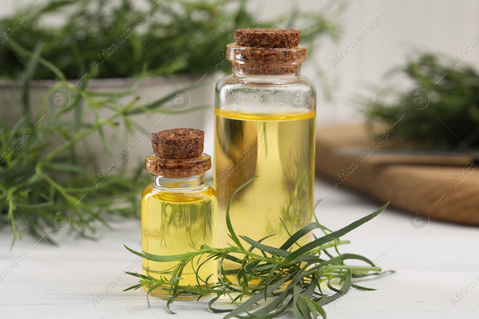 Photo of Bottles of essential oil and fresh tarragon leaves on white wooden table