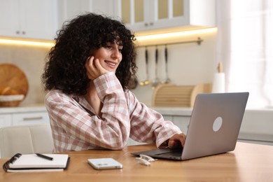 Photo of Beautiful young woman in stylish pyjama using laptop at wooden table in kitchen