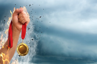 Image of Winner raising hand with gold medal from fire flames up to stormy sky, closeup. Space for text