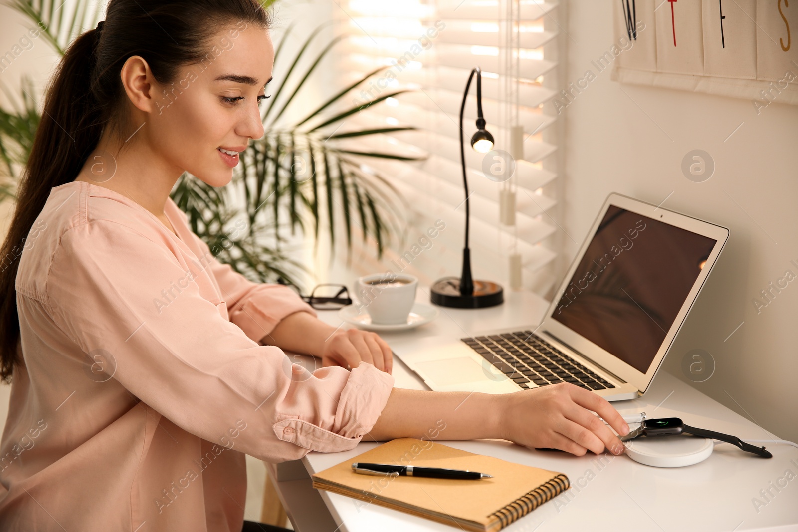 Photo of Woman putting smartwatch onto wireless charger at white table. Modern workplace accessory