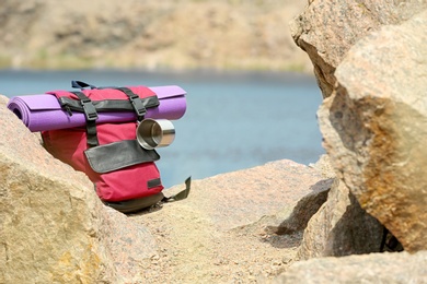 Photo of Camping backpack with mat and mug on stones near river. Space for text