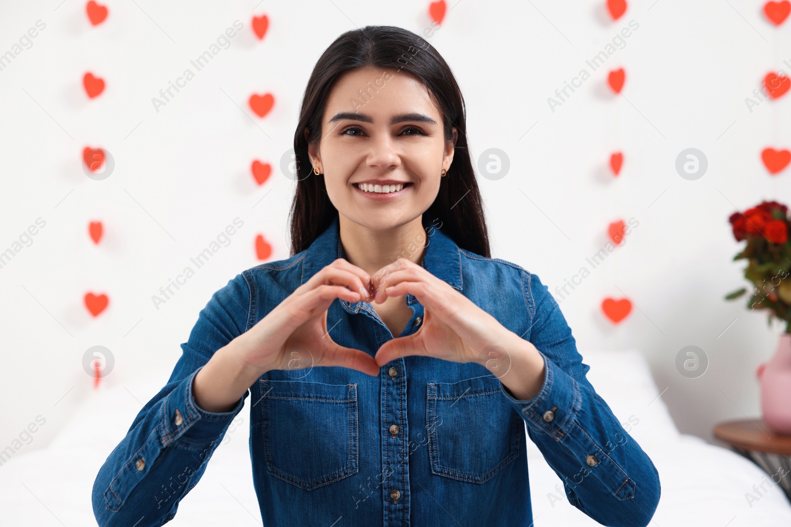 Photo of Beautiful young woman making heart with hands indoors, view from camera. Valentine's day celebration in long distance relationship