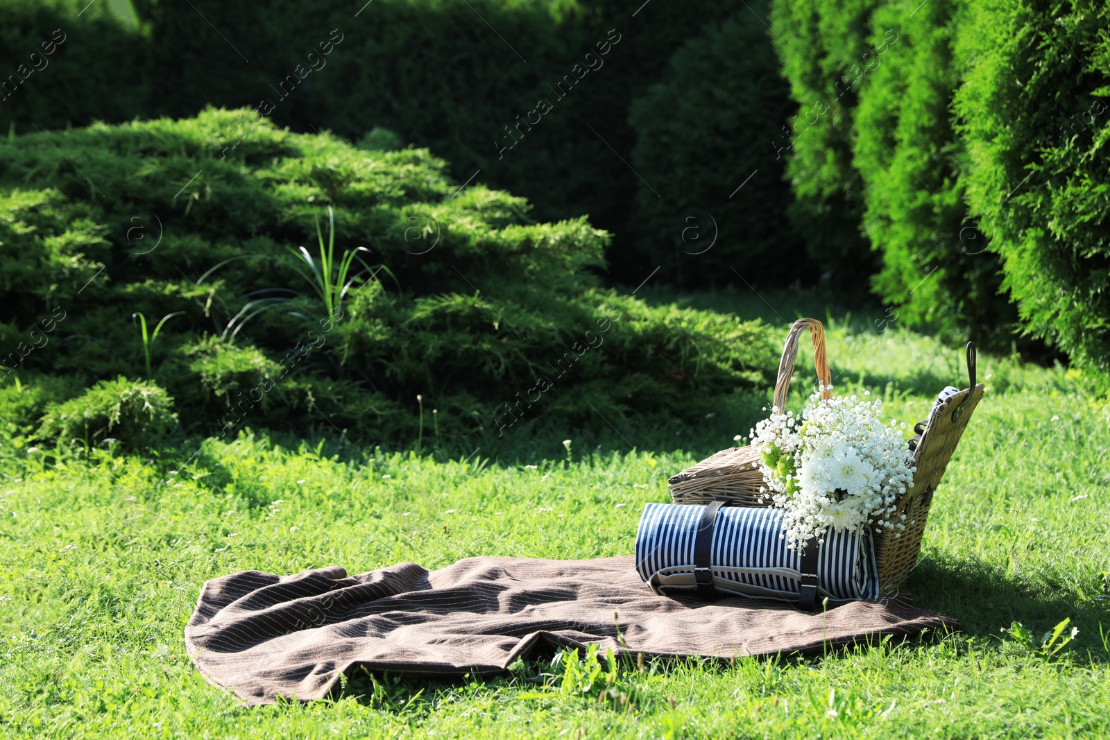 Photo of Picnic blanket with beautiful flowers, rolled mat and basket in garden on sunny day
