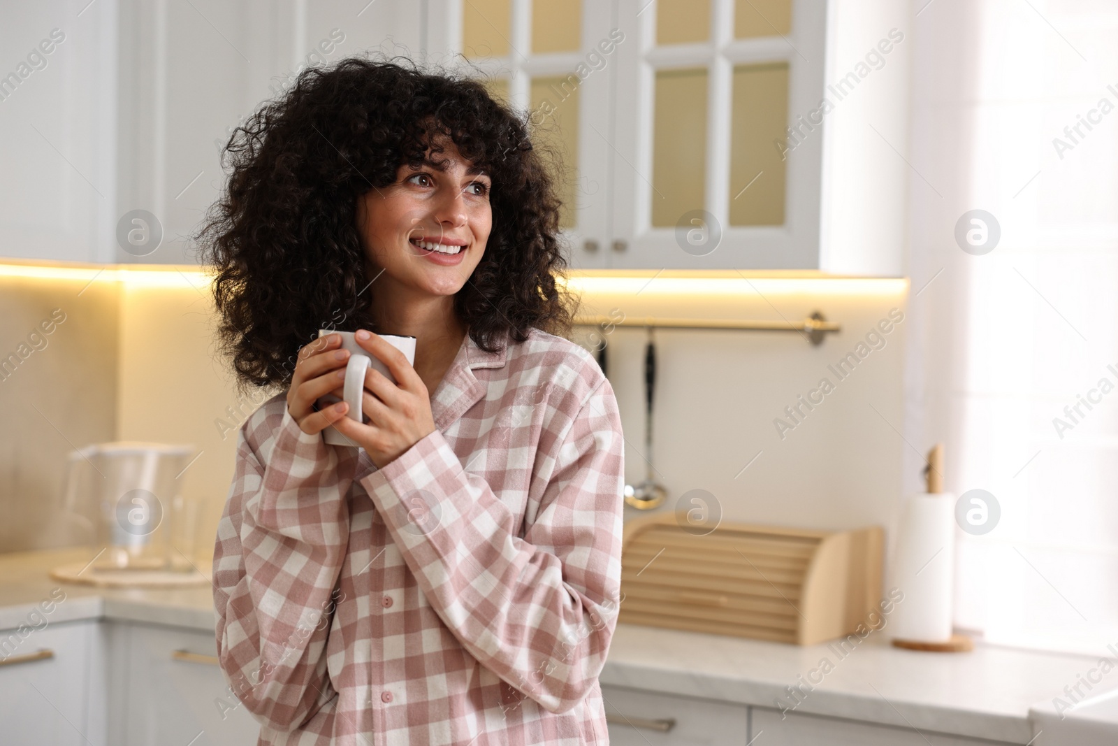 Photo of Beautiful young woman in stylish pyjama with cup of drink in kitchen, space for text