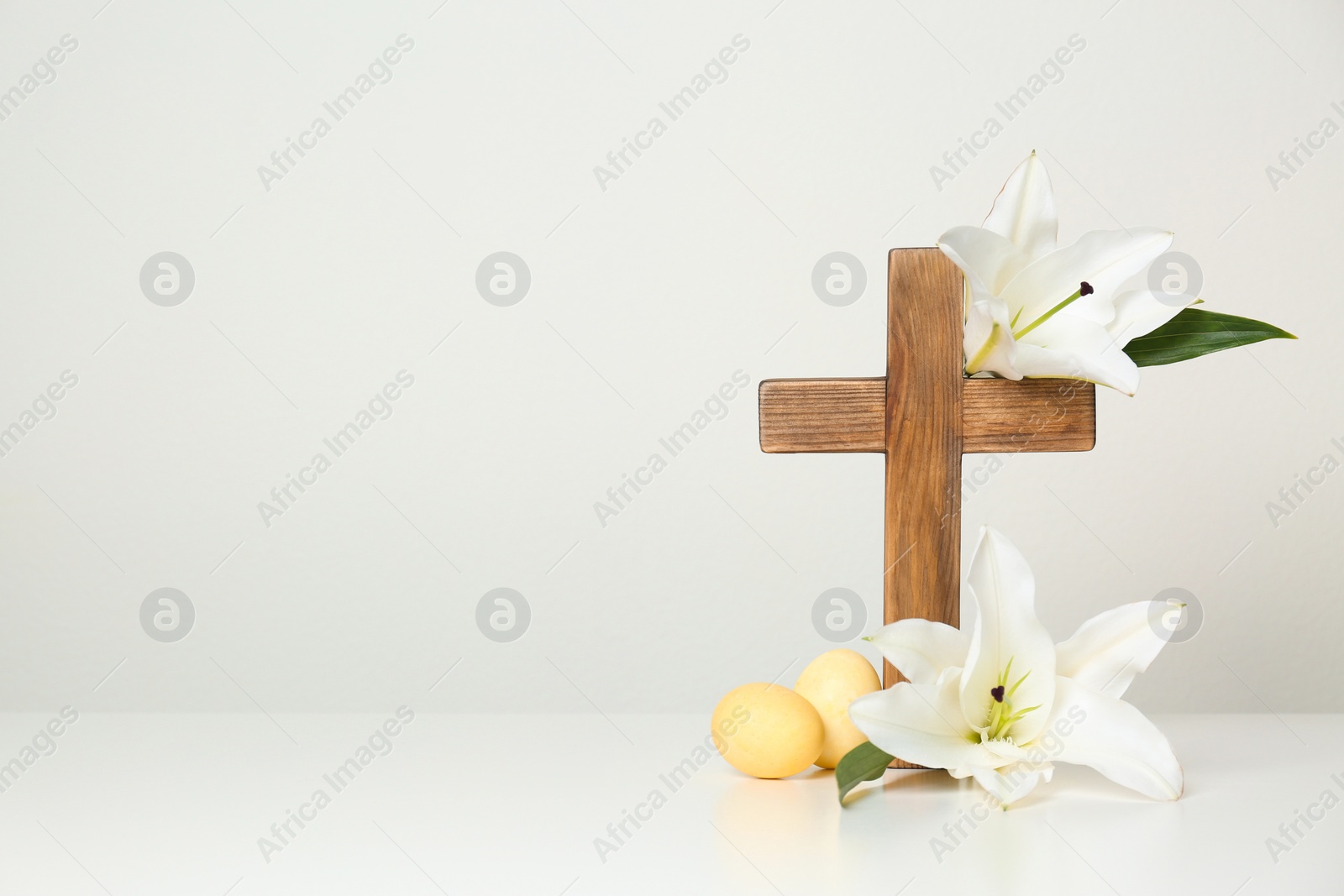 Photo of Wooden cross, Easter eggs and blossom lilies on table against light background, space for text