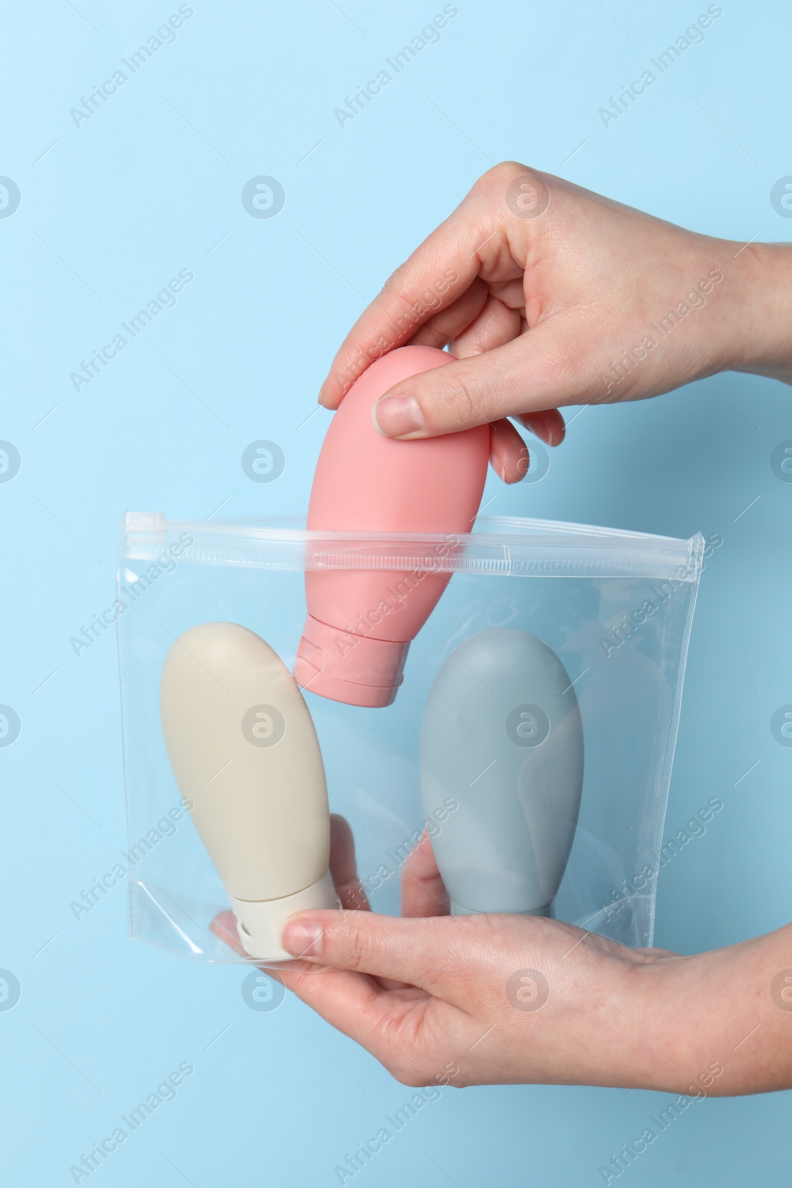 Photo of Cosmetic travel kit. Woman putting small bottle with personal care product into plastic bag against light blue background, closeup