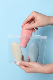 Cosmetic travel kit. Woman putting small bottle with personal care product into plastic bag against light blue background, closeup