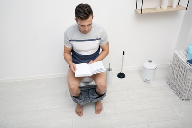 Photo of Young man with book sitting on toilet bowl in bathroom
