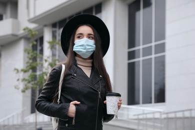 Photo of Young woman in medical face mask with cup of coffee walking outdoors. Personal protection during COVID-19 pandemic