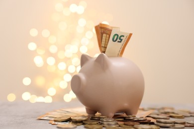 Photo of Piggy bank with euro banknote and coins on grey table against blurred lights