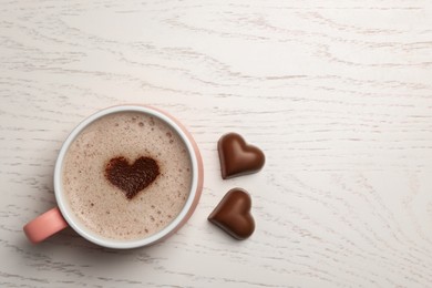 Cup of aromatic coffee with heart shaped decoration and chocolate candies on white wooden table, flat lay. Space for text