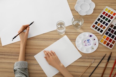 Photo of Woman painting with watercolor on blank paper at wooden table, top view