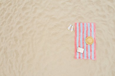 Striped beach towel, book, straw hat and flip flops on sand, aerial view. Space for text