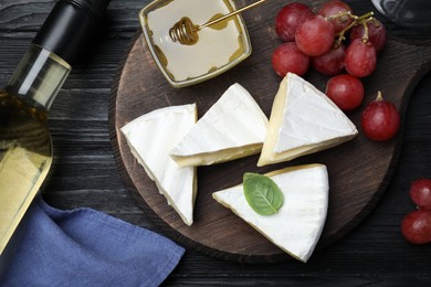 Photo of Tasty brie cheese with basil, grapes, honey and wine on black wooden table, flat lay