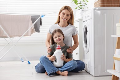 Photo of Mother and daughter sitting on floor near washing machine and holding fabric softener in bathroom