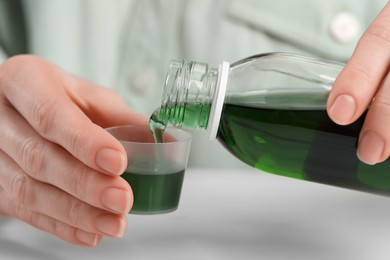 Photo of Woman pouring syrup from bottle into measuring cup at table, closeup. Cold medicine