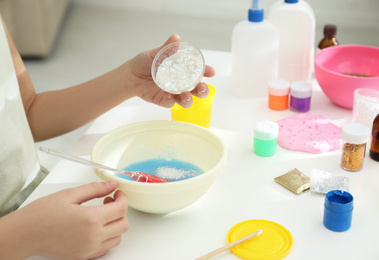 Photo of Little girl adding sparkles into homemade slime toy at table, closeup of hands