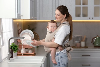 Mother holding her child in sling (baby carrier) while washing plates in kitchen