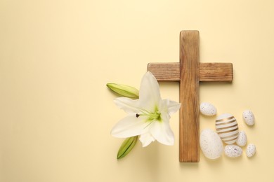 Wooden cross, painted Easter eggs and lily flowers on pale yellow background, flat lay. Space for text