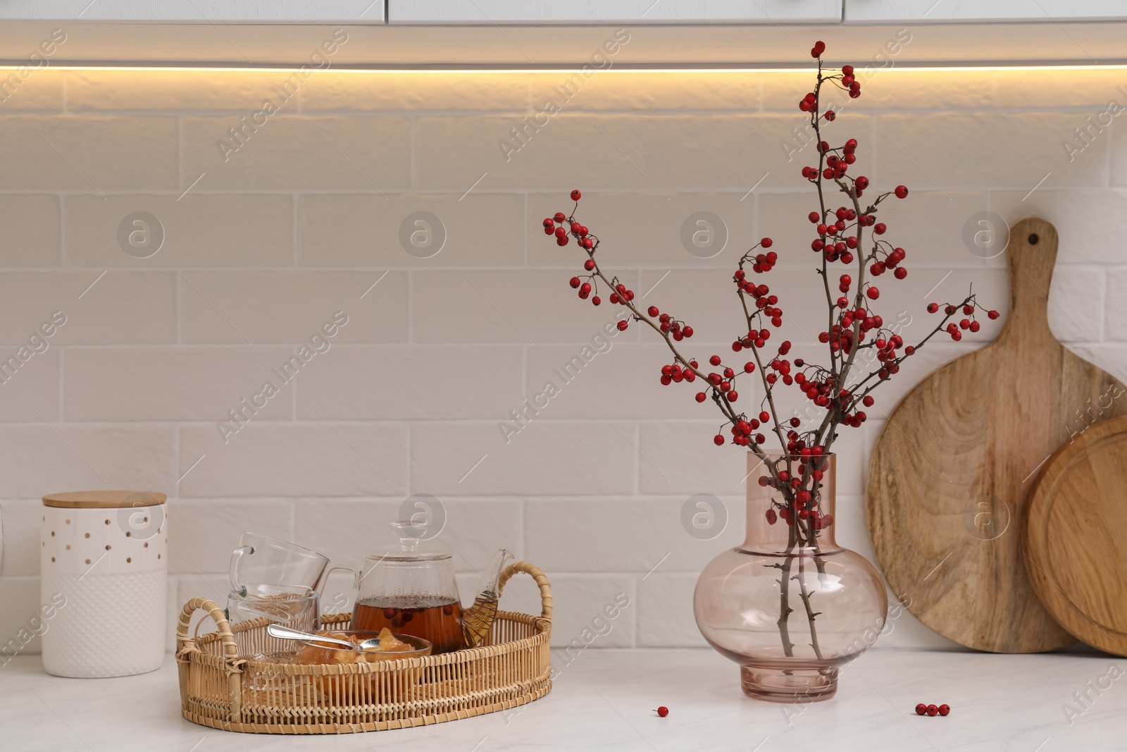 Photo of Hawthorn branches with red berries in vase and aromatic tea on white table indoors