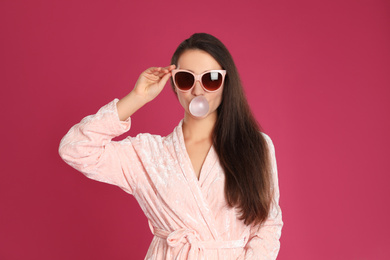 Photo of Young woman in bathrobe and sunglasses blowing chewing gum on crimson background