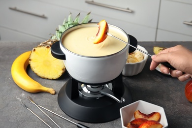 Photo of Woman dipping peach into pot with white chocolate fondue on table, closeup