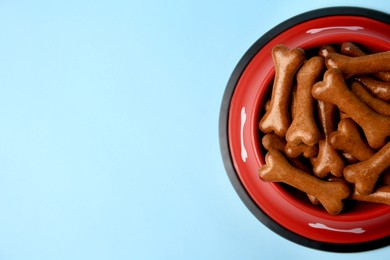 Photo of Red bowl with bone shaped dog cookies on light blue background, top view. Space for text