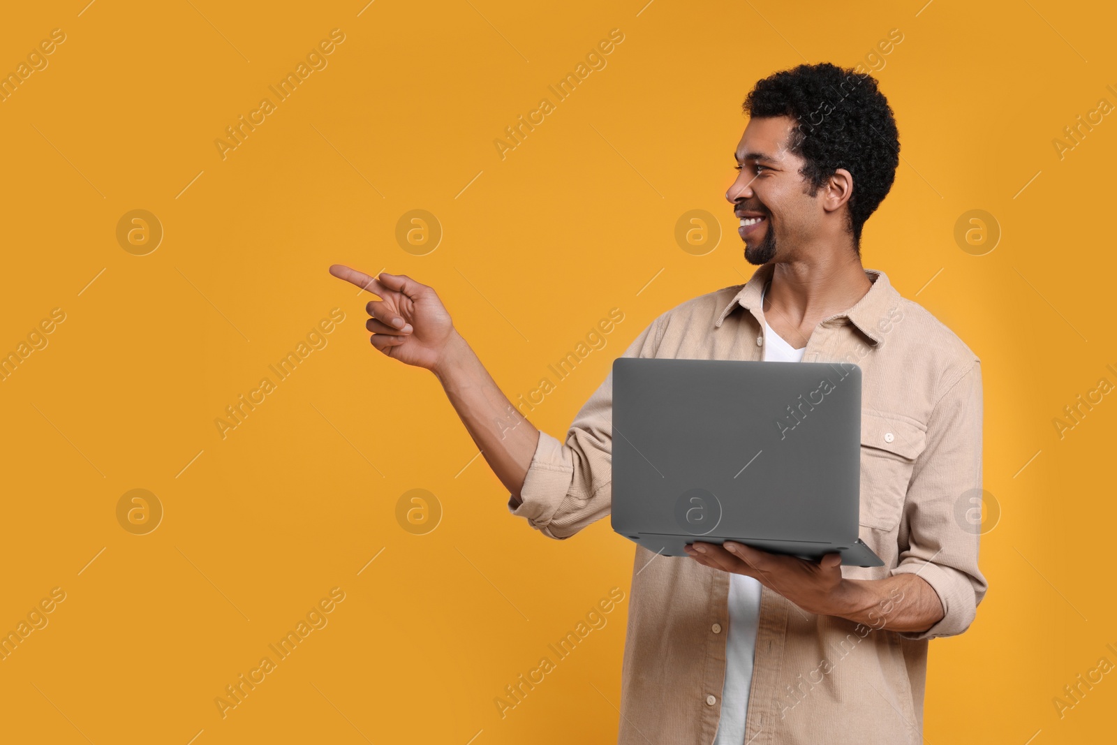 Photo of Happy man with laptop pointing at something on orange background. Space for text