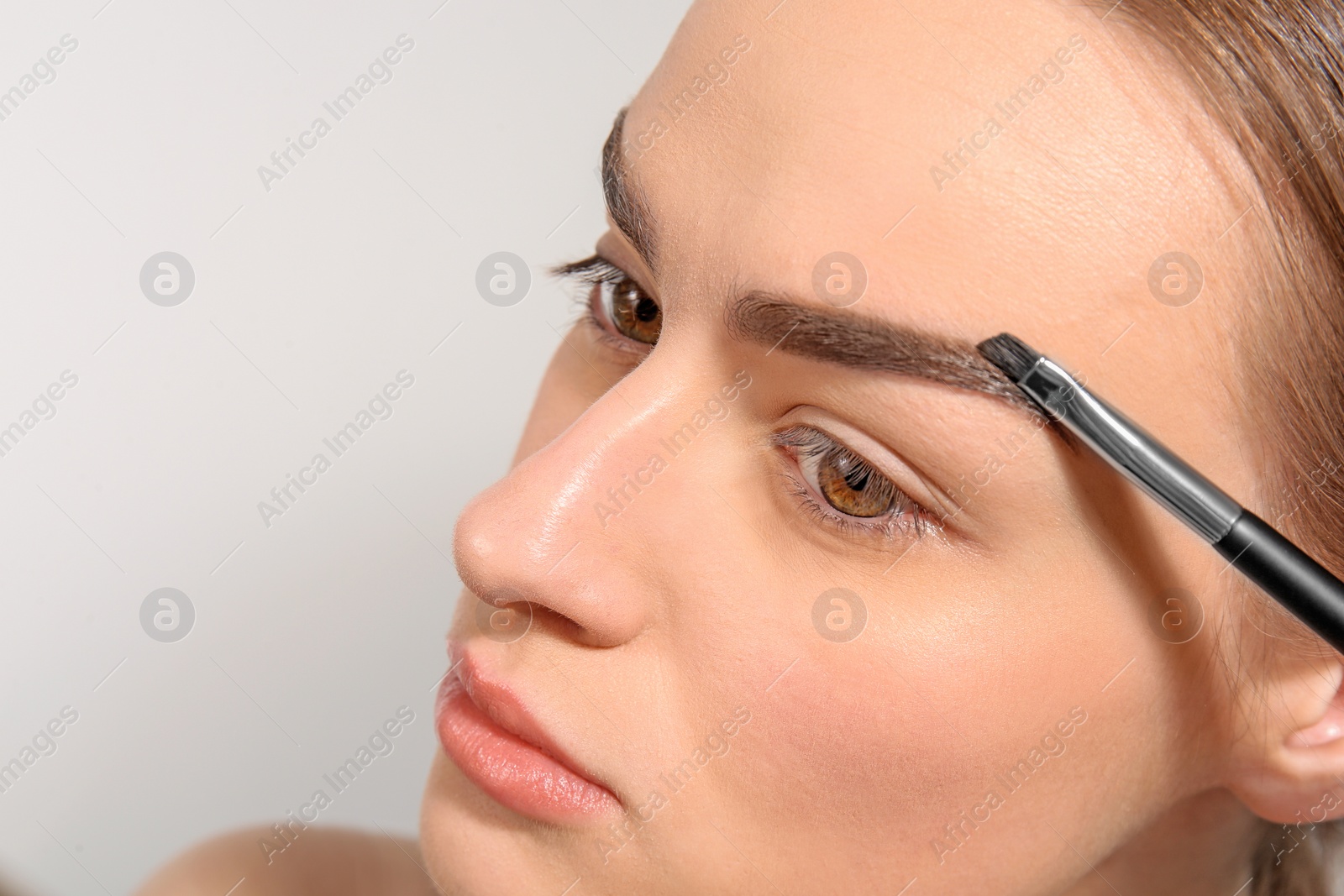 Photo of Young woman correcting shape of eyebrow with brush on light background