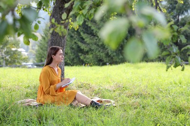 Photo of Beautiful young woman drawing with pencil in notepad on green grass near tree