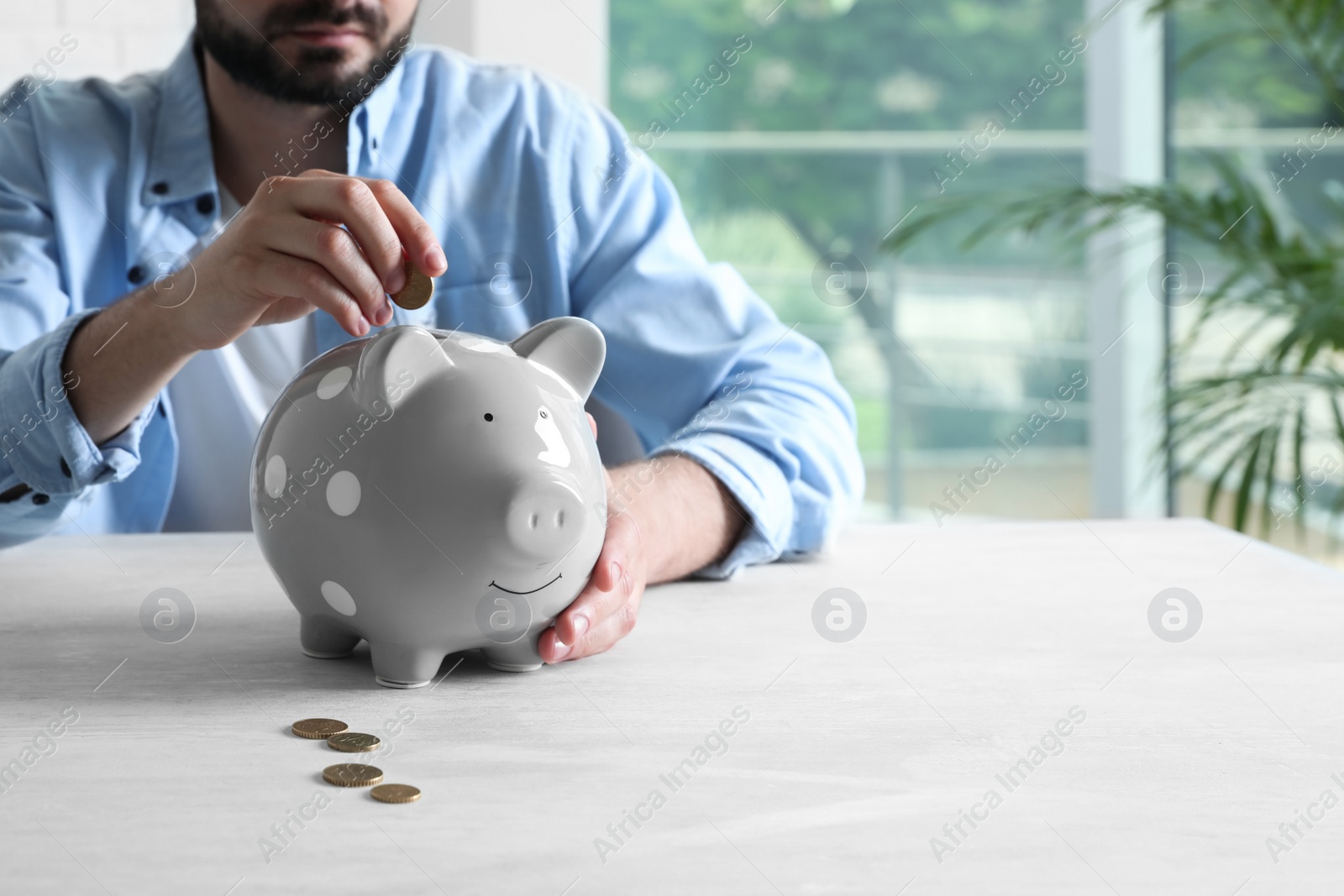 Photo of Man putting coin into piggy bank at white table indoors, closeup