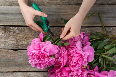 Photo of Woman trimming beautiful pink peonies with secateurs at wooden table, top view