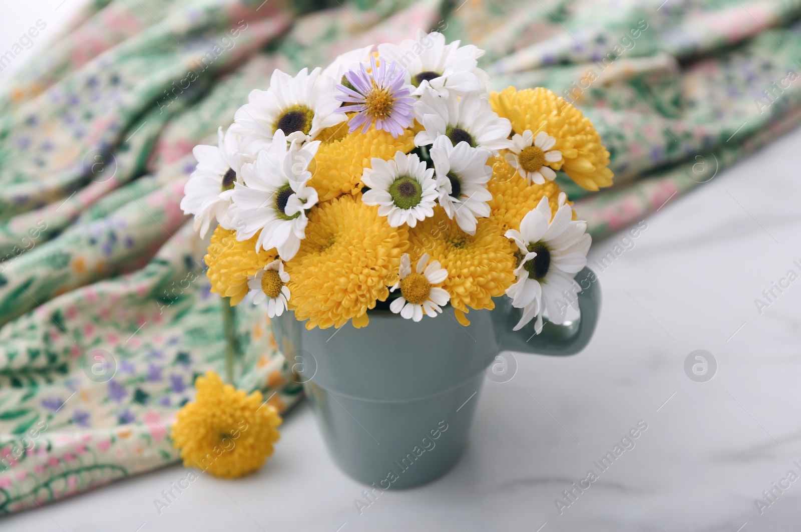 Photo of Beautiful bright flowers in cup and fabric on white marble table, closeup