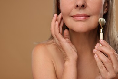 Photo of Woman massaging her face with jade roller on brown background, closeup