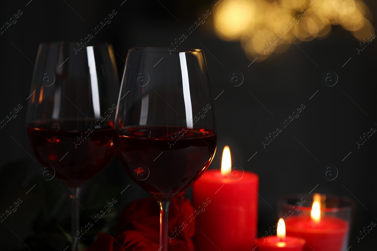 Photo of Glasses of red wine, burning candles and rose flowers against blurred lights, space for text. Romantic atmosphere