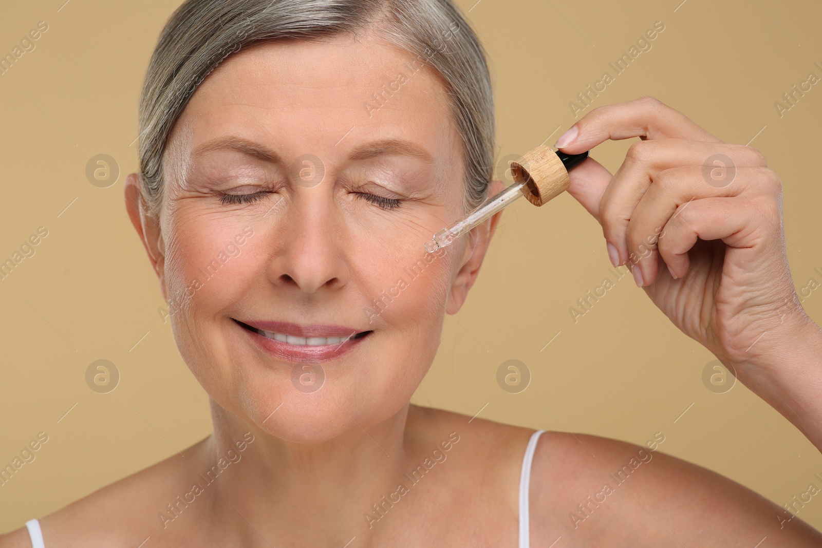 Photo of Senior woman applying cosmetic product on her aging skin against beige background. Rejuvenation treatment