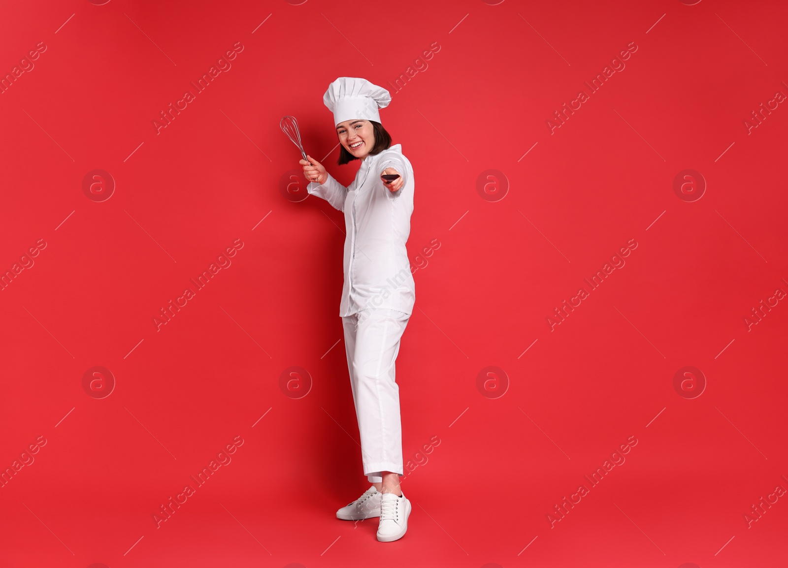 Photo of Happy confectioner holding whisk and spatula on red background