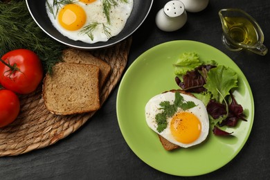 Photo of Frying pan and plate with tasty fried eggs, salad and products on black table, flat lay