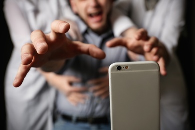 Photo of Emotional man reaching for smartphone while medical workers holding him back on black background, closeup. Addiction concept