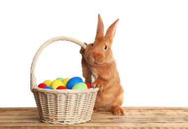Photo of Adorable furry Easter bunny near wicker basket with dyed eggs on wooden table against white background